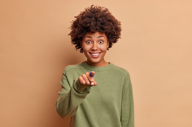 Uma modelo feminina de aparência agradável e sorridente aponta o dedo da frente diretamente para a frente e vê algo engraçado na frente de seus sorrisos com dentes brancos vestidos casualmente isolados sobre a parede marrom