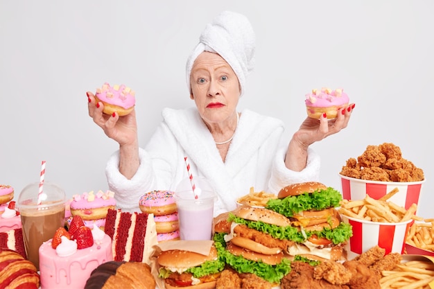 Foto grátis uma mulher aposentada e hesitante e surpresa segurando duas deliciosas rosquinhas com glacê cercada por alimentos nocivos com alto teor calórico