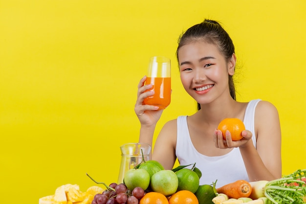 Uma mulher asiática vestindo uma camiseta branca. segurando um copo de suco de laranja com a mão direita a mão esquerda se movia e havia muitas frutas na mesa.
