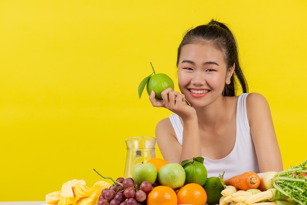 Uma mulher asiática vestindo uma camiseta branca. Segurando uma faixa com a mão direita E na mesa há muitas frutas diferentes.