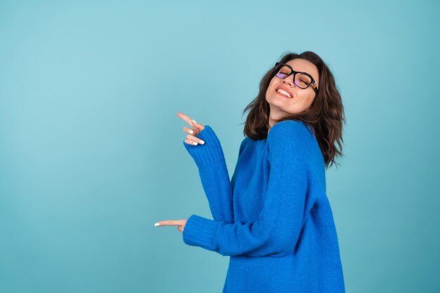 Uma mulher de suéter de malha azul, cabelo curto encaracolado, óculos nos olhos, sorri alegremente, apontando para um espaço vazio à esquerda