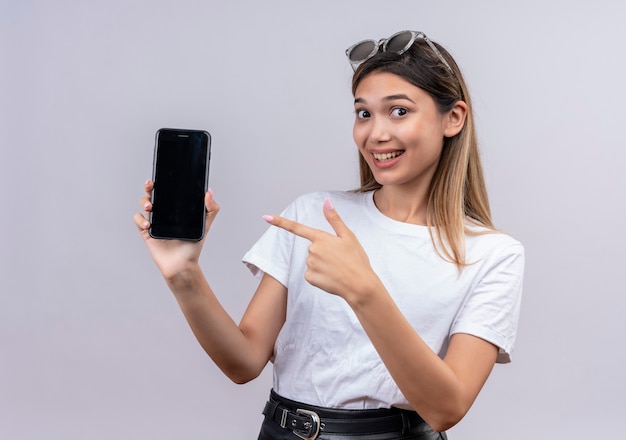 Uma mulher jovem e positiva em uma camiseta branca com óculos de sol sorrindo enquanto mostra o espaço em branco do telefone celular enquanto olha para uma parede branca