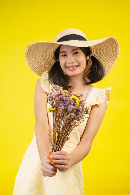 Uma mulher linda e feliz usando um chapéu grande e segurando um buquê de flores secas em um amarelo.