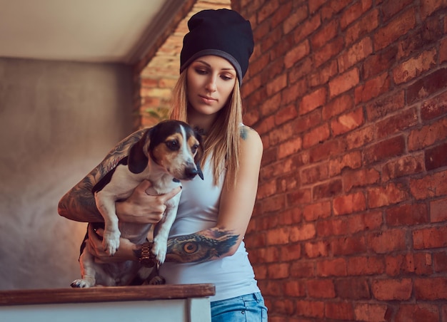 Uma mulher loira tatuada elegante em t-shirt e jeans segura um cachorro fofo.