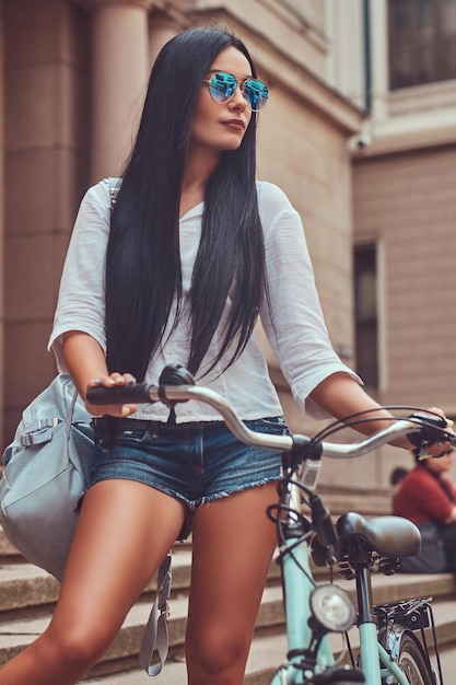 Foto grátis uma mulher morena sexy vestindo blusa e shorts jeans em óculos de sol, posando com uma bicicleta perto de um prédio antigo na cidade.