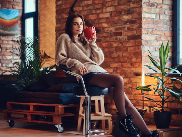 Uma mulher morena vestida com uma jaqueta marrom longo pescoço bebe café quente em uma sala com interior loft.