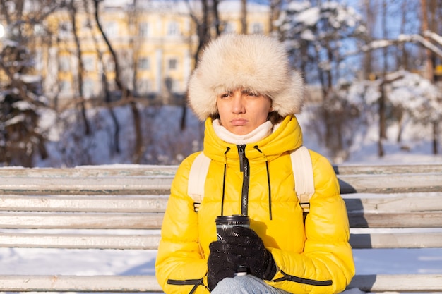Uma mulher no inverno com roupas quentes em um parque coberto de neve em um dia ensolarado está sentada em um banco e está congelando de frio, é infeliz no inverno, segura o café sozinha