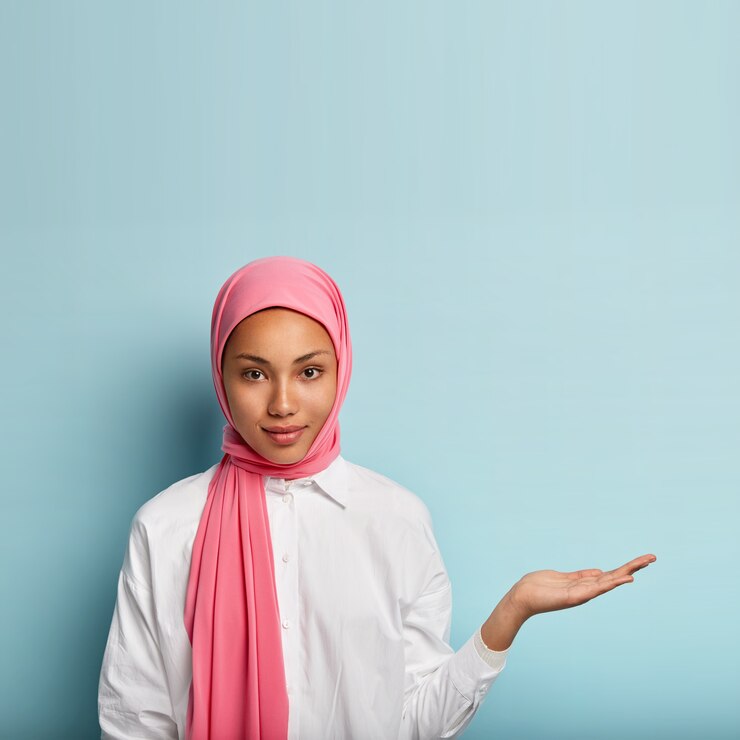 Foto grátis uma mulher religiosa muçulmana confiante levanta a palma da mão sobre o espaço em branco, segura um objeto invisível, usa um lenço de seda rosa na cabeça, promove um item, fica de pé contra a parede azul, vestido com uma camisa branca