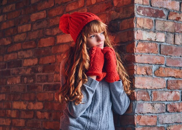 Uma mulher ruiva atraente vestida com um chapéu quente e luvas posando sobre a parede de um tijolo vermelho.