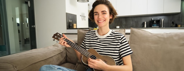 Foto grátis uma rapariga bonita a tocar ukulele no sofá a cantar a sua canção favorita e a pegar acordes na guitarra pequena