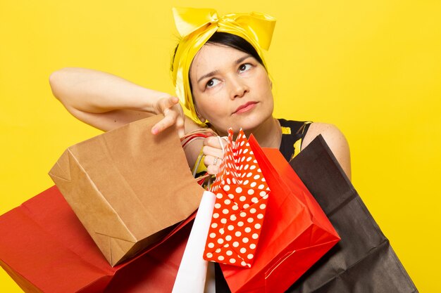 Uma senhora de vista frontal em vestido amarelo-preto flor projetado com bandagem amarela na cabeça posando segurando pacotes de compras no amarelo