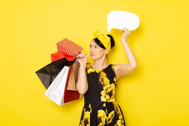Uma senhora de vista frontal em vestido amarelo-preto flor projetado com bandagem amarela na cabeça segurando pacotes de compras sinal branco sobre o amarelo