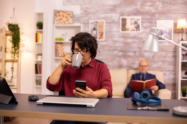 Uma senhora idosa na casa dos 60 anos usando tecnologia moderna em um ambiente aconchegante enquanto o marido lê ao fundo