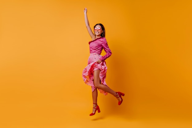 Uma senhora satisfeita e animada com sapatos com salto urbano firme está pulando em roupas de seda rosa claro. Retrato de corpo inteiro de uma menina com cabelo macio e macio se movendo em um quarto laranja