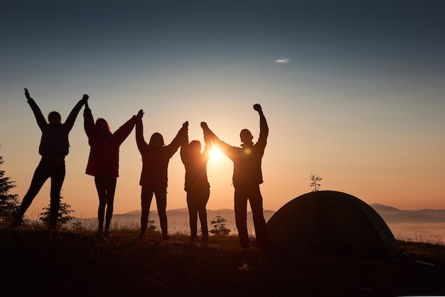 Uma silhueta de um grupo de pessoas se diverte no topo da montanha perto da tenda durante o pôr do sol.