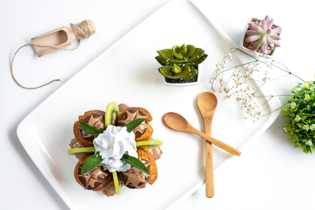 Uma sobremesa choco vista de cima com kiwis fatiados e creme dentro de uma mesa branca, juntamente com colheres de madeira e plantas na mesa branca, frutas doces exóticas