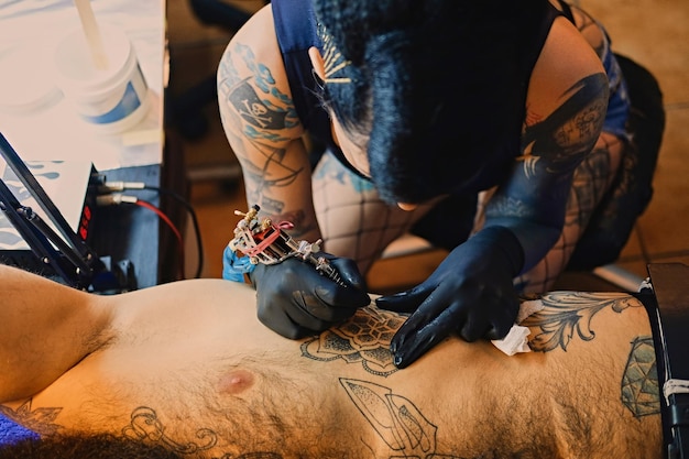 Uma tatuadora profissional faz tatuagem preto e branco no torso masculino barbudo em um salão.