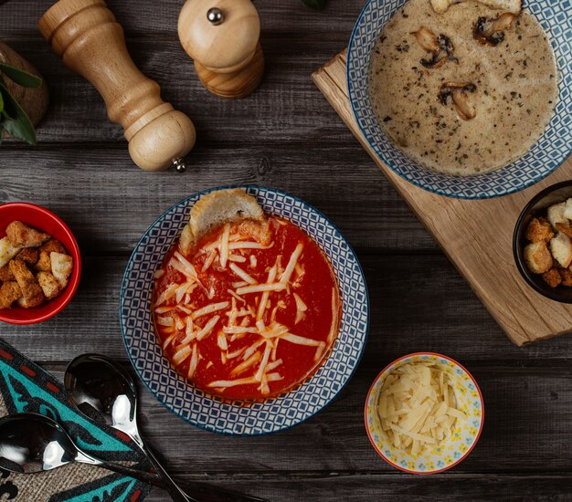 Uma tigela azul de sopa de tomate com queijo parmesão picado no topo e sopa de cogumelos ao redor