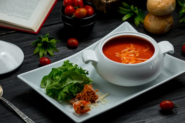 Uma tigela branca de sopa de tomate com parmesão picado e salada verde.