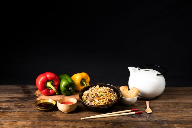 Uma tigela de carne delicioso teriyaki com macarrão udon; molho de soja; pimentão e primavera papel com pauzinhos e colher na mesa de madeira