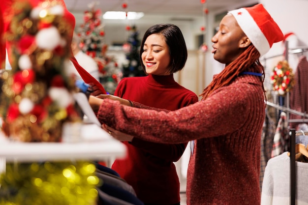 Foto grátis uma vendedora a ajudar um cliente asiático a encontrar a medida certa, a recomendar roupas e acessórios para os preparativos do jantar de natal no centro comercial. mulheres diversas a discutir a qualidade dos tecidos.