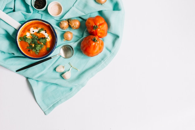 Uma visão aérea da sopa de tomate com ingredientes na toalha de mesa azul contra um fundo branco