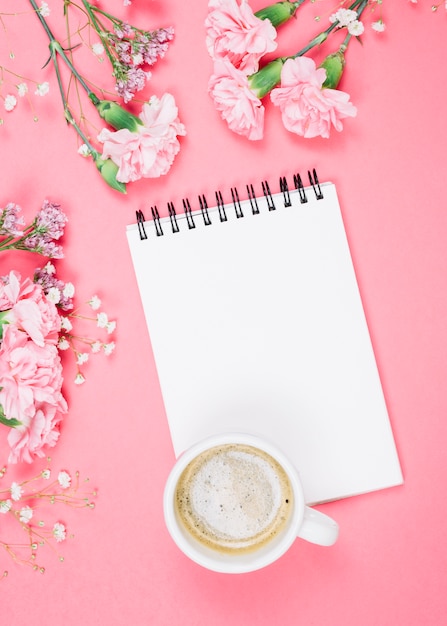 Uma visão aérea da xícara de café no bloco de notas em branco com cravos; gipsila; limonium flores sobre fundo rosa