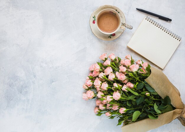 Uma visão aérea de buquê de flores cor de rosa; xícara de café; bloco de notas em espiral e caneta em pano de fundo concreto