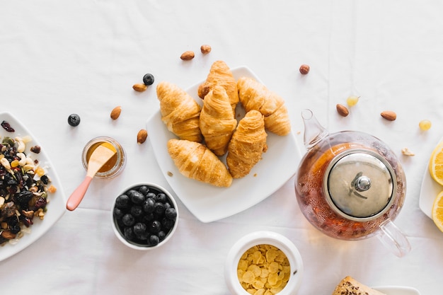 Uma visão aérea de croissant assado; frutas; chá e dryfruits na toalha de mesa branca