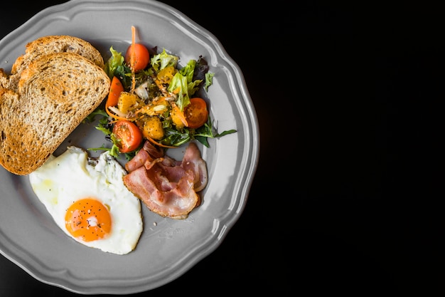 Uma visão aérea de torrada; bacon; salada e ovos fritos na chapa cinza sobre fundo preto