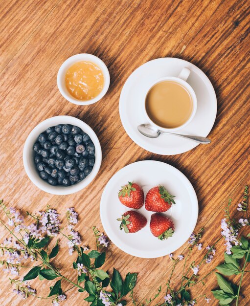 Uma visão elevada de mirtilo; geléia; xícara de morango e café em flores contra o pano de fundo de madeira