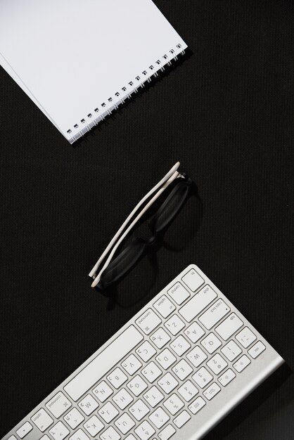 Uma visão elevada do bloco de notas em espiral; óculos e teclado na mesa preta