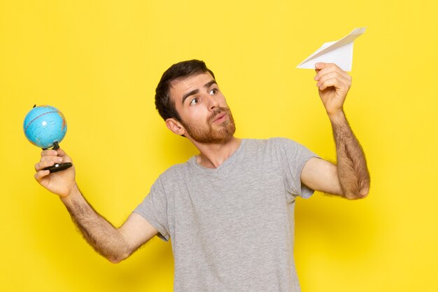 Uma visão frontal de um jovem do sexo masculino com uma camiseta cinza segurando um avião de papel branco e um pequeno globo no modelo de cor amarela do homem