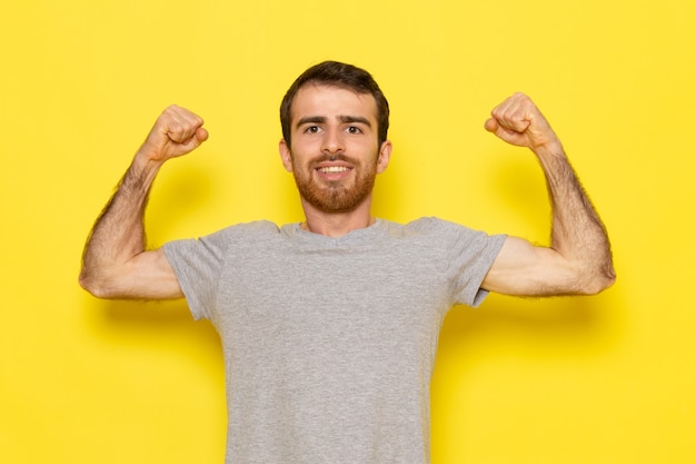 Uma visão frontal jovem do sexo masculino em uma camiseta cinza sorrindo e flexionando na parede amarela homem cor modelo emoção roupas