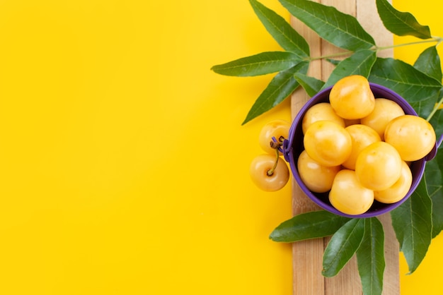 Uma vista de cima cerejas amarelas suaves e doces com folhas verdes em uma mesa amarela, cor de frutas cereja doce de verão