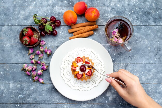 Uma vista de cima, um delicioso pequeno bolo com creme e frutas frescas fatiadas, juntamente com chá de canela no bolo de mesa cinza-azulado.