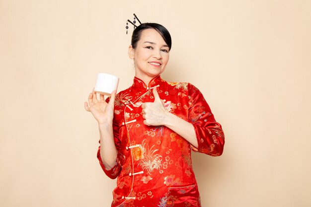 Uma vista frontal bela gueixa japonesa no tradicional vestido japonês vermelho com varas de cabelo posando usando lata de creme sobre a cerimônia de creme de fundo japão
