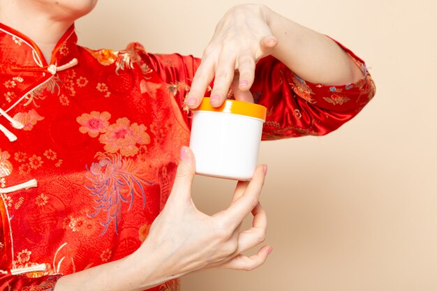 Uma vista frontal bela gueixa japonesa no tradicional vestido vermelho japonês com varas de cabelo posando segurando creme pode sobre o fundo creme cerimônia japão