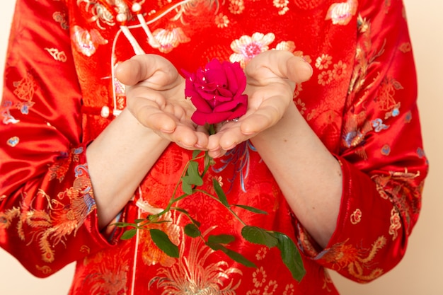 Uma vista frontal bela gueixa japonesa no vestido tradicional vermelho japonês com varas de cabelo posando segurando rosa vermelha elegante na cerimônia de creme de fundo divertido Japão Oriental