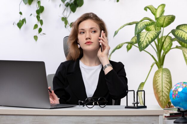 Uma vista frontal bela moça de camisa branca e jaqueta preta, usando o laptop na frente da mesa, falando ao telefone com as folhas penduradas