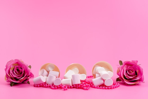 Uma vista frontal com marshmallow dentro de pacotes de papel junto com rosas rosa isoladas em uma mesa rosa, confeitaria açucarada