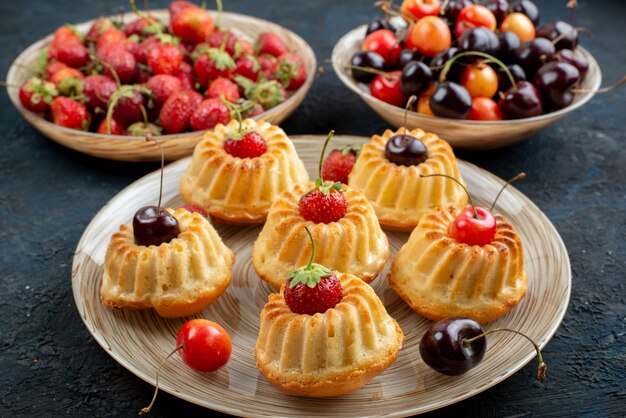 Uma vista frontal de biscoitos deliciosos com morangos e cerejas vermelhas dentro do prato na mesa escura