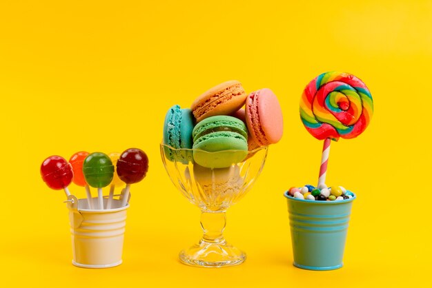 Uma vista frontal de macarons franceses junto com balas e pirulitos dentro de baldes em doces doces de confeitaria amarelos