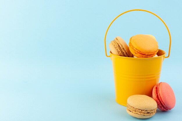 Uma vista frontal de macarons franceses multicoloridos dentro de um balde na mesa azul