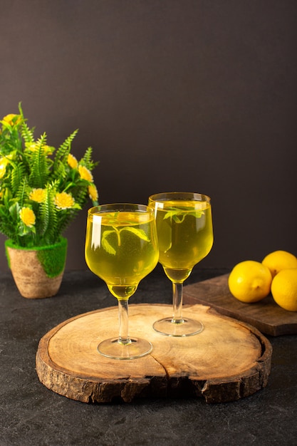 Uma vista frontal de óculos com suco de suco de limão dentro de vidros transparentes ao longo de limão inteiro e flores na mesa de madeira marrom e bebida de limão coquetel de fundo cinza