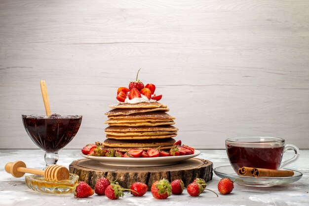 Uma vista frontal deliciosas panquecas redondas com chá de creme e morangos vermelhos no bolo de mesa de madeira