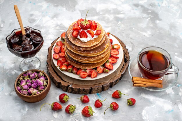 Uma vista frontal deliciosas panquecas redondas com creme e chá de morangos vermelhos no bolo de mesa de madeira