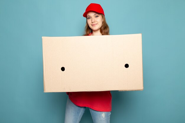 Uma vista frontal feminino atraente correio em camisa polo vermelha boné vermelho e calça jeans segurando a caixa posando sorrindo sobre o trabalho de serviço de alimentação de fundo azul