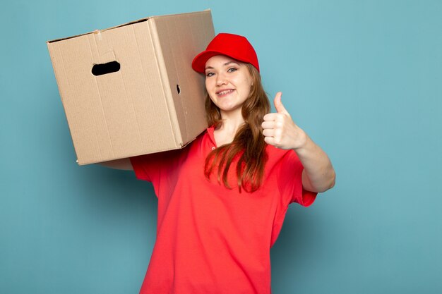 Uma vista frontal feminino atraente correio em camisa polo vermelha boné vermelho e calça jeans segurando a caixa posando sorrindo sobre o trabalho de serviço de alimentação de fundo azul