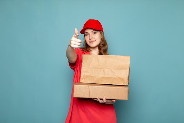 Uma vista frontal feminino atraente correio em camisa polo vermelha boné vermelho segurando pacotes marrons sorrindo posando no trabalho de serviço de alimentação de fundo azul